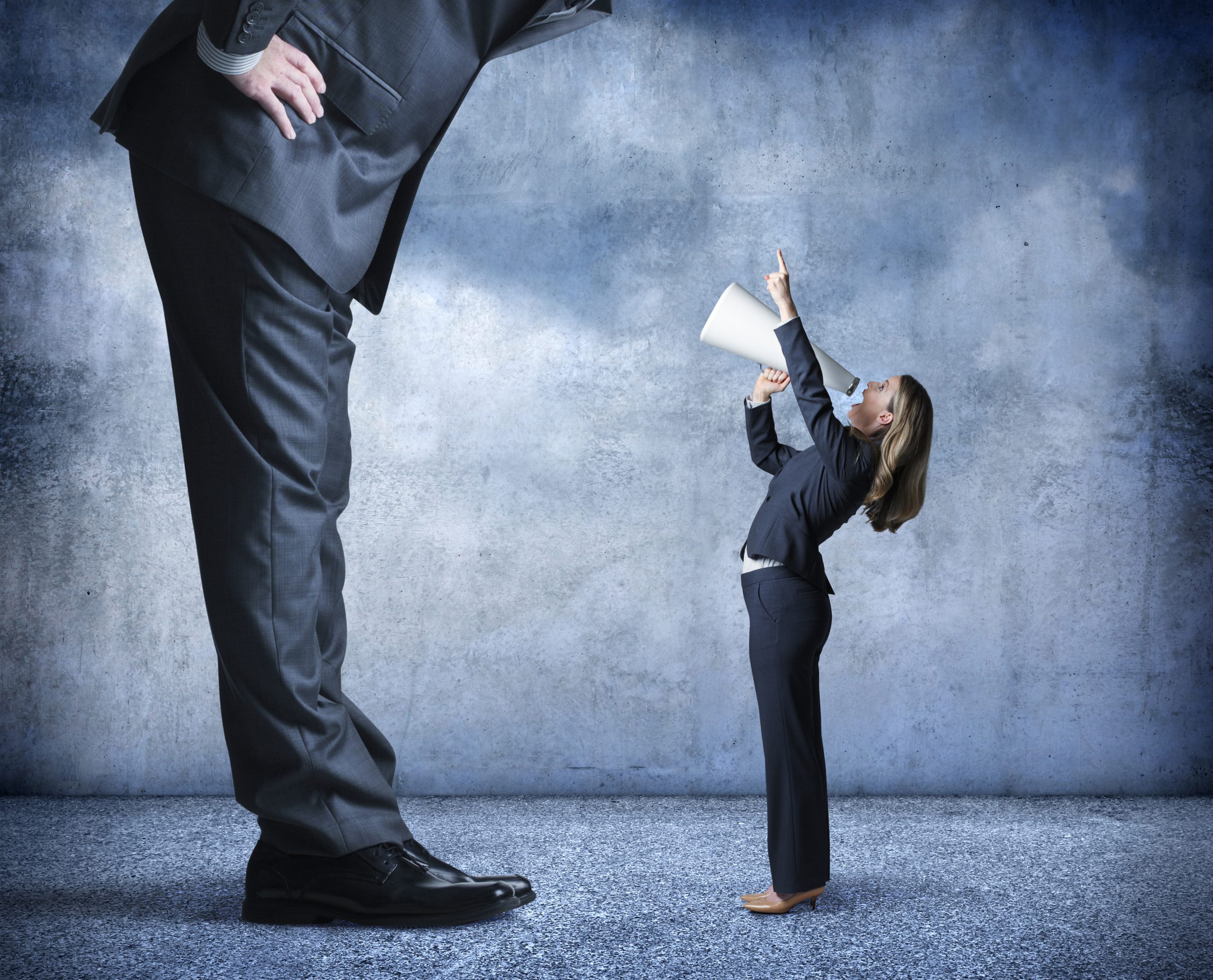 A businesswoman shouts through a megaphone and points up towards a much larger businessman who is standing over her.
